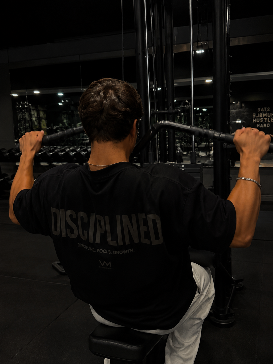 A man in a gym, viewed from behind, wearing a black t-shirt with 'DISCIPLINED' printed in gray across the back while pulling a cable on a machine.
