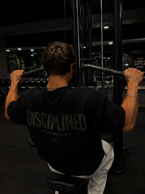 A man in a gym, viewed from behind, wearing a black t-shirt with 'DISCIPLINED' printed in gray across the back while pulling a cable on a machine.