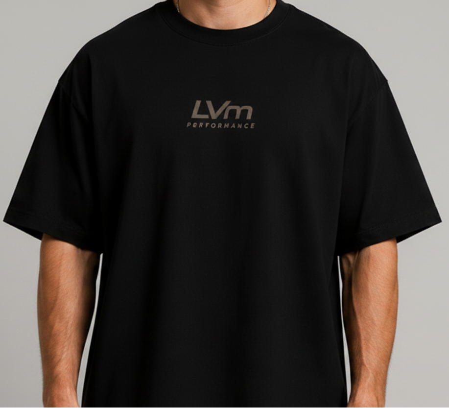 A man wearing an oversized black t-shirt with a metallic gold LVm Performance logo on the chest, photographed against a neutral gray background.